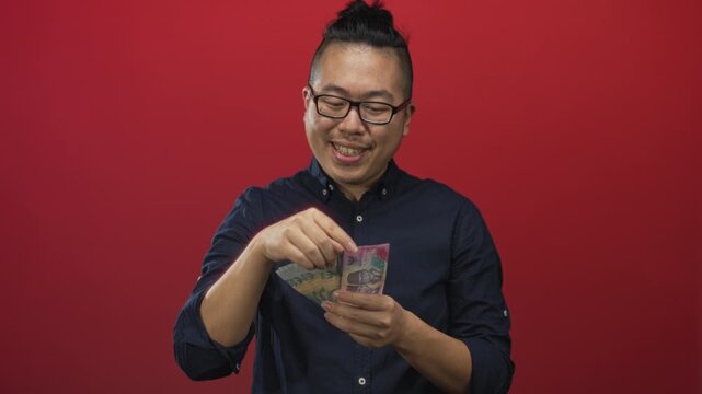 Indonesian man smiling and counting rupiah banknotes with visible hands in red studio; savings contentment.