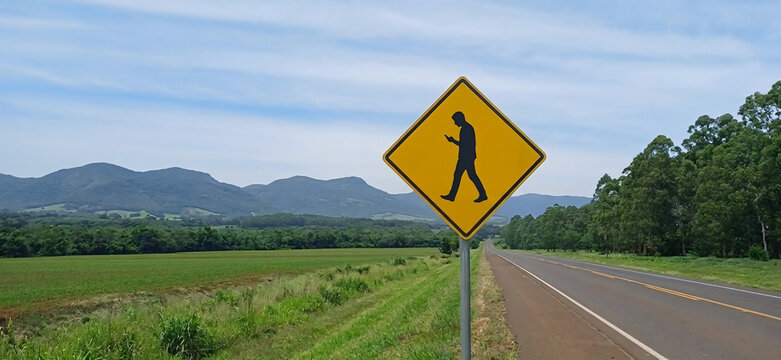 Warnschild f&uuml;r abgelenkte Fu&szlig;g&auml;nger mit Smartphone an einer Landstra&szlig;e vor Berglandschaft