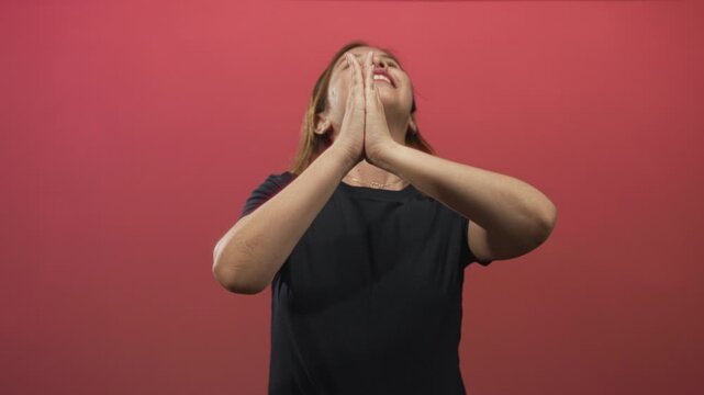 Young caucasian woman wearing black tshirt with praying hands clasped and looking up in red studio; hope faith gratitude.