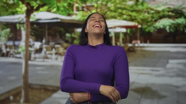 Woman in a purple top covering her mouth with hand while laughing on a leafy city street under cafe umbrellas; happiness.