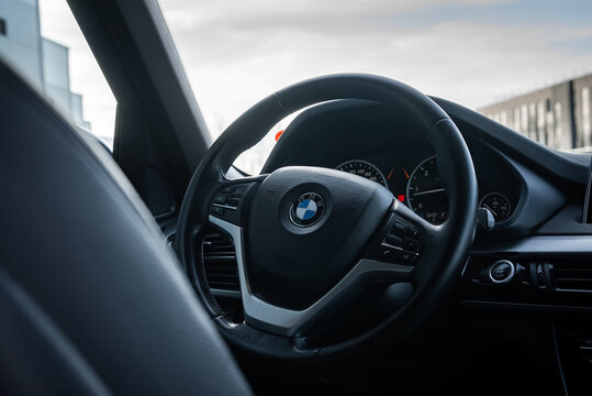 BMW X5 interior shows roundel on wheel, multifunction controls, and analog gauges, parked on a modern street with contemporary buildings in soft daytime light.