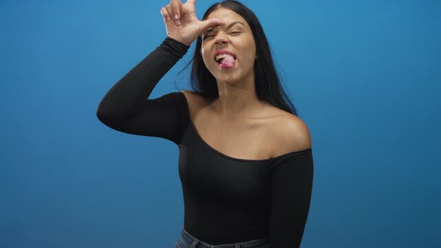 Woman with long black hair making loser sign on forehead and sticking tongue out in blue studio; defiance.