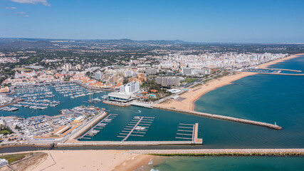People visit Vilamoura marina in Portugal's Algarve region, enjoying boats and beach while the city skyline stands tall in the distance under clear blue sky Aerial © Sergo