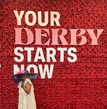 At Churchill Downs, home of the Kentucky Derby, A Woman in Wide-Brim Hat Smiles in Front of Rose Wall with 'Your Derby Starts Now' Sign