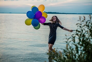 Happy young woman splashing water in lake with colorful balloons at sunset © Bohdan