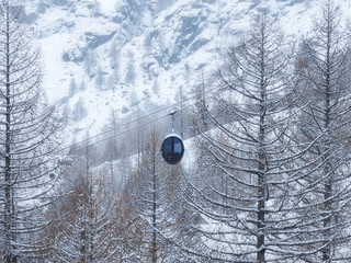A single enclosed gondola glides above frosted larches at San Domenico in the Italian Alps, with rugged alpine cliffs rising behind, mid winter light and quiet mood. © Aerial Film Studio