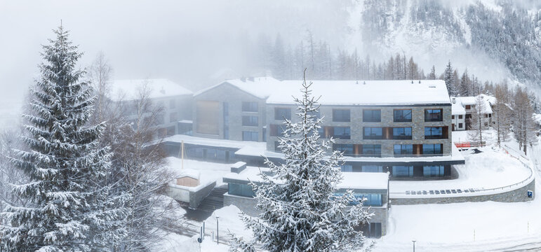 San Domenico ski resort in the Italian Alps shows a modern loft hotel, skiers' terrain, access paths, fir trees, and misted cliff lined peaks in winter conditions.