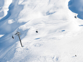Aerial scene at San Domenico in the Italian Alps shows chairlifts, gondola cabins, a lift tower, skiers descending wide pistes, and broad snowfields with faint tracks. © Aerial Film Studio