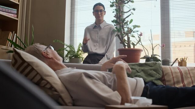 Rack focus shot of focused middle-aged Caucasian female therapist standing by window in modern office and listening attentively to mature male patient lying on couch and sharing emotions