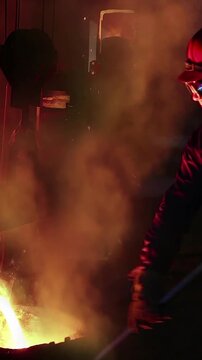 Liquid metal in the foundry, melting iron in furnace, steel mill. Worker with goggles and helmet watching and controlling iron smelting in furnaces