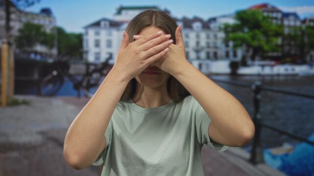 Caucasian teenage girl with brown hair covers eyes with hands while standing on street by canal railing under blue sky; shyness.