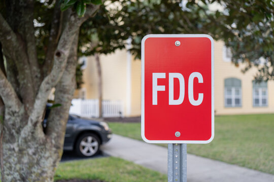 Vertical outdoor scene of a reflective FDC sign in front of a suburban property, framed by tree bark, grass, and softly blurred housing backdrop
