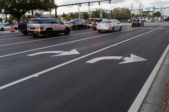 Cars moving and waiting through a large signalized intersection, with turn lane arrows, smooth black pavement, curb edge, and overhead traffic lights