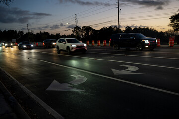 Fototapeta premium Evening road scene in America with reflective black asphalt, curved lane arrows, moving cars, and a long line of orange safety barrels at dusk