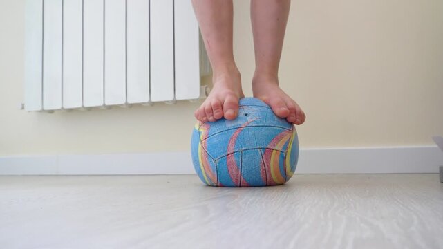 The child's feet are on a bright soft ball in a room with warm light. The scene conveys the mood of play, balance, and activity at home. Foot massage.