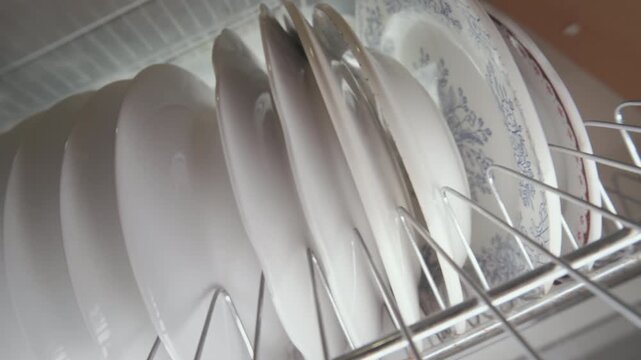 Close-Up Low Angle View: White Ceramic Plates Standing on Metal Dish Rack Inside Kitchen Cabinet | Clean Dishes Stored on Shelf | Home Organization and Kitchen Storage Concept