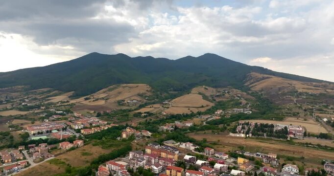 Aerial panorama of Vulture Mount, in Basilicata, Italy. It's a sprawling mountain range under a dramatic, cloudy sky. Below the peaks lies a patchwork of golden fields and small clusters of houses.