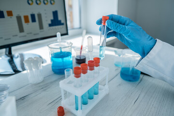 Fototapeta premium Close up of a gloved hand handling a test tube filled with blue chemical solution in a laboratory. Advanced technology and scientific research tools are present on the white worktable