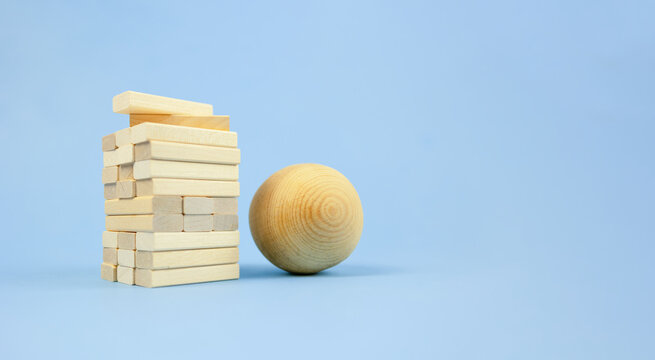 wooden ball leaning against tall block tower on blue backdrop, visual metaphor for risk management in business workshop, dynamic tension and texture, studio lighting emphasizes grain