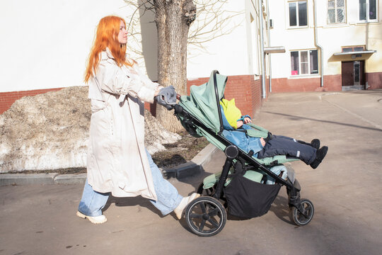 Mature redhead mother walking with toddler in stroller on city street in spring.