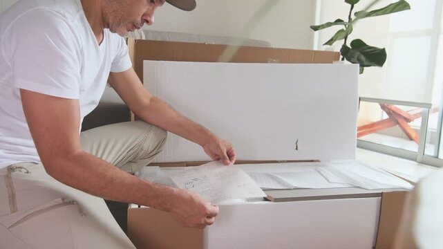 Focused man carefully unpacking a cardboard box and reading the instruction manual for new furniture