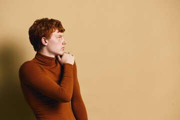 Profile portrait of young man with red hair wearing brown turtleneck sweater, looking thoughtful against beige background. Casual style with natural light and contemplative mood. © SHOTPRIME STUDIO