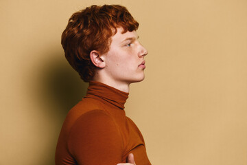Profile portrait of young man with red hair wearing brown turtleneck sweater looking forward against plain yellow background, side view with serious expression and casual style. © SHOTPRIME STUDIO