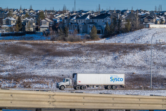 Calgary, Alberta, Canada. March 13, 2026. A Sysco semi-trailer truck travels along a highway with a suburban residential neighborhood in the background.