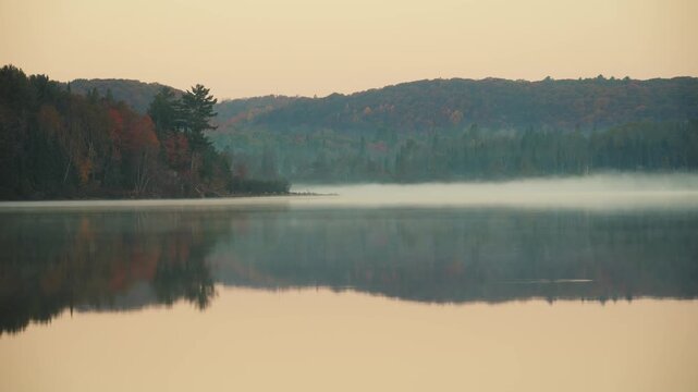 Sunrise timelapse over calm lake and autumn forest, morning mist and fog moving across water as sunlight hits colorful fall foliage and hills