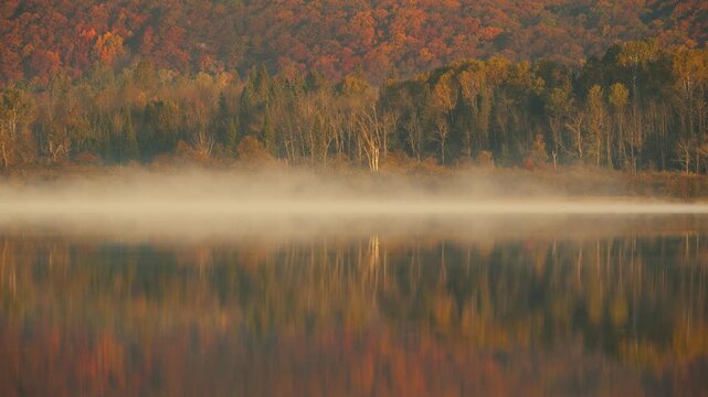 Morning fog rising over calm lake at sunrise with autumn forest reflection, ethereal mist moving across still water surface, cinematic fall nature timelapse
