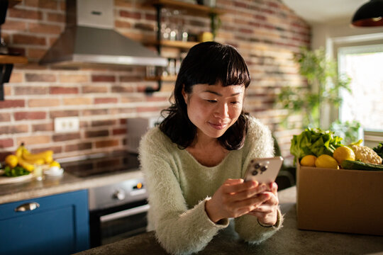 Smiling woman using smartphone in home kitchen with fresh grocery delivery