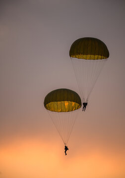 Two combat paratroopers deploying by parachute during an air show demonstration. Silhouettes of troopers descending with glowing parachute canopies against a sunset sky