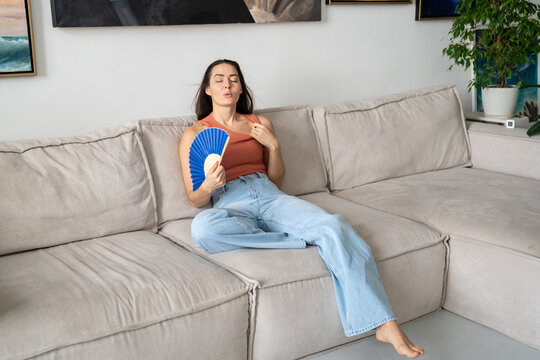 Woman waving paper fan cooling herself during summer heatwave, sitting on couch at home dealing with heatstroke. Female enjoying air from hand fan, feeling stress and dehydration, unhealthy heat.