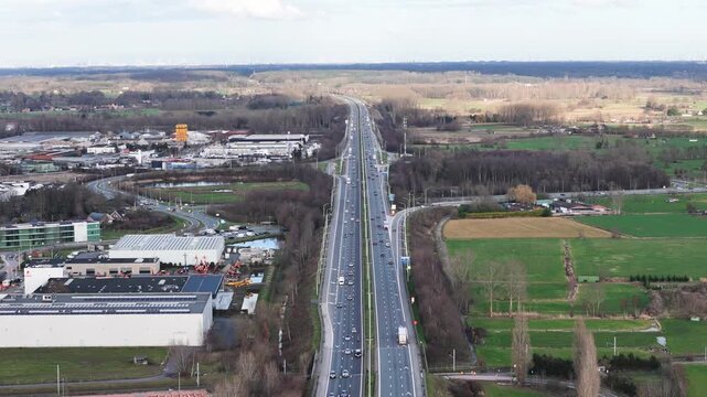 high altitude multilane highway stretching horizon traffic industrial buildings one green fields houses woods other toward long steady side drone view multi-lane 