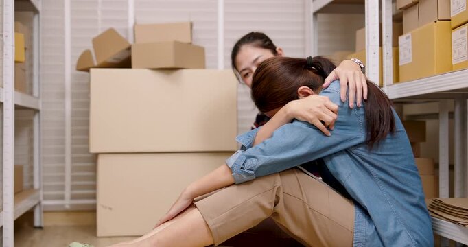Asian women partners supporting small business teamwork, comforting exhausted partner inside home warehouse, emotional during e-commerce operation, managing parcel box workload pressure together