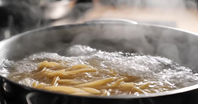 Boiling pasta in a pot on a stove, steam rising, with kitchen utensils in the background