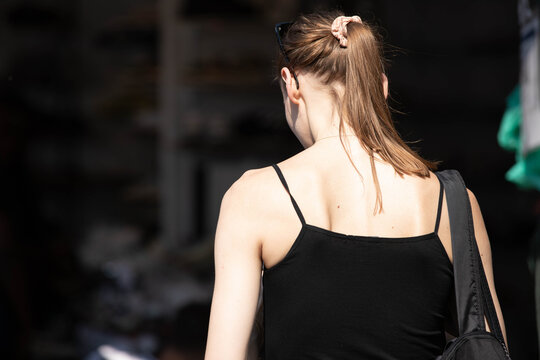 Female traveler in black tank top shopping in market, pedestrian walking in harsh sunlight, skin texture, rear view, at Chatuchak Market.