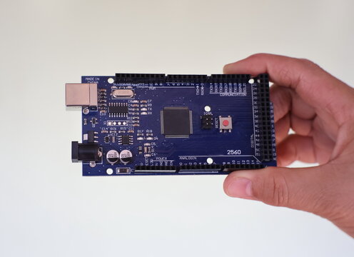 A close-up image of a hand holding a blue Arduino Mega 2560 Rev3 microcontroller board against a white background.