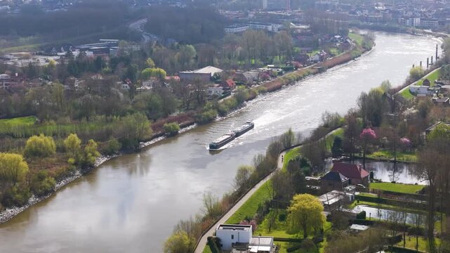 cargo vessel cruising sunlit river bend bordered by leafy neighborhoods garden embankments curving roadway leading hazy urban skyline riverside area to aerial shot transportation waterway 
