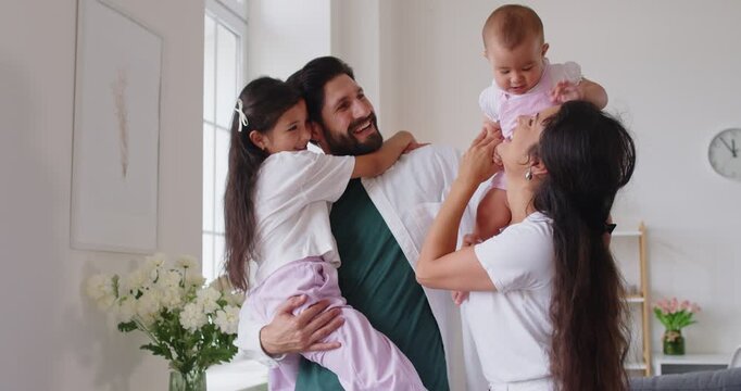 Family parents baby daughter home bonding together. Portrait of playful moment in a bright room as mom and dad hold kids, smiling in natural light. Love, care and happy family concept.