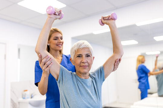 Senior woman exercising with dumbbells during physical therapy with a doctor. Shoulder strengthening and geriatric rehabilitation concept. Elderly patient lifting weights for muscle health in clinic.