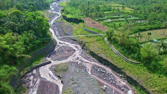 Aerial drone footage of a long thin metallic bridge, crossing a wide rocky river, with little water streaming down, with forest around, in Semeru lava area, Lumajang regency, Java island, Indonesia