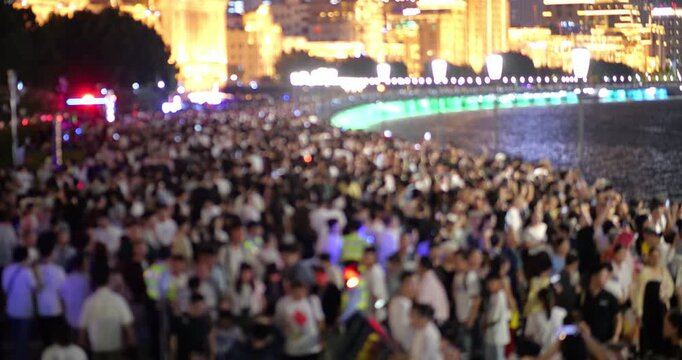 Blurred view of thousands of people walking along waterfront in continuous motion, diminishing perspective of curved embankment. Warm and bright glow of illuminated buildings on background.