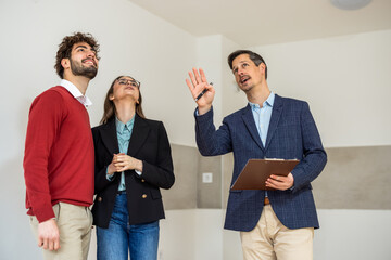 Real estate agent pointing at the ceiling while showing an empty apartment to a young couple.