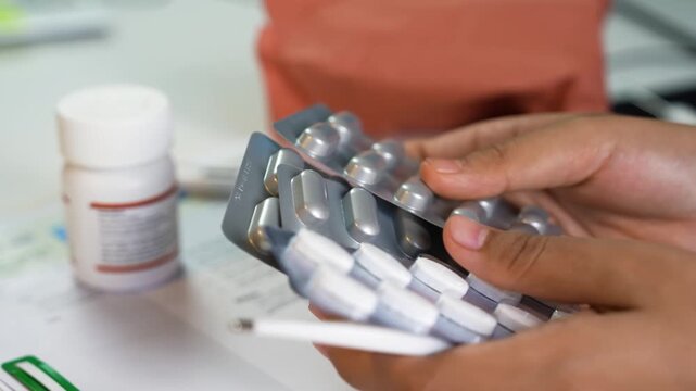 Hands holding blister packs of pills with pill bottle and pen on table, medical medication context