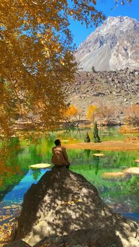 Traveler Sitting on Rock by Emerald Mountain Lake With Autumn Trees enjoying solitude in Naltar Valley Pakistan