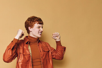 Young redhead woman celebrating success with fists raised and happy face against beige background....