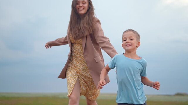 Happy mother and son are running in green field together. Little boy runs fast while woman chases him outdoors. Active family plays in nature park. Smiling parent and child enjoy fun run in grass.