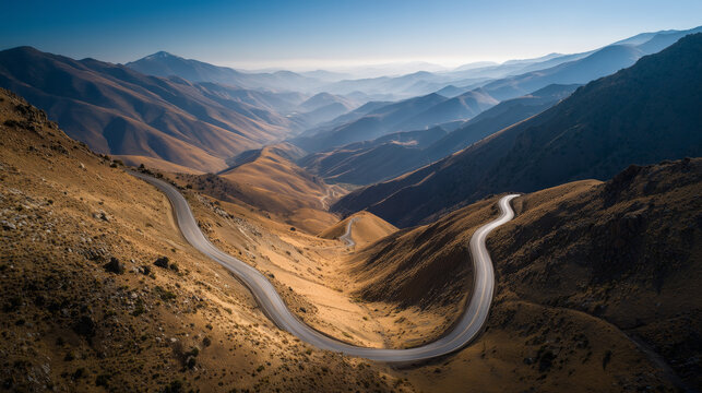 Winding mountain road through a vast, sunlit valley with layered hills and a clear blue sky.