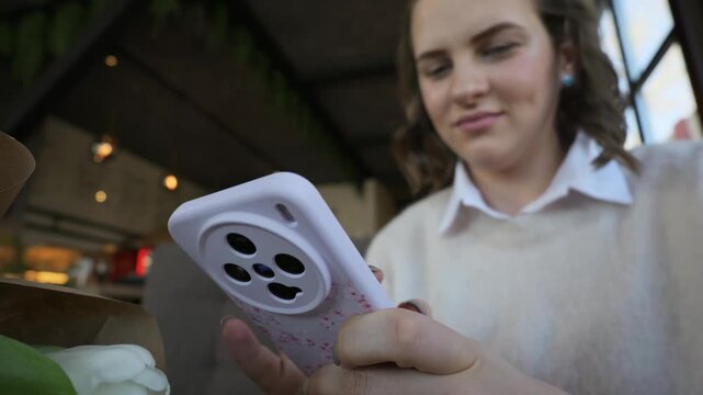 Female hands type on a smartphone with a prominent white camera case inside a cozy cafe. Concept of daily digital communication, social media browsing, and modern lifestyle.